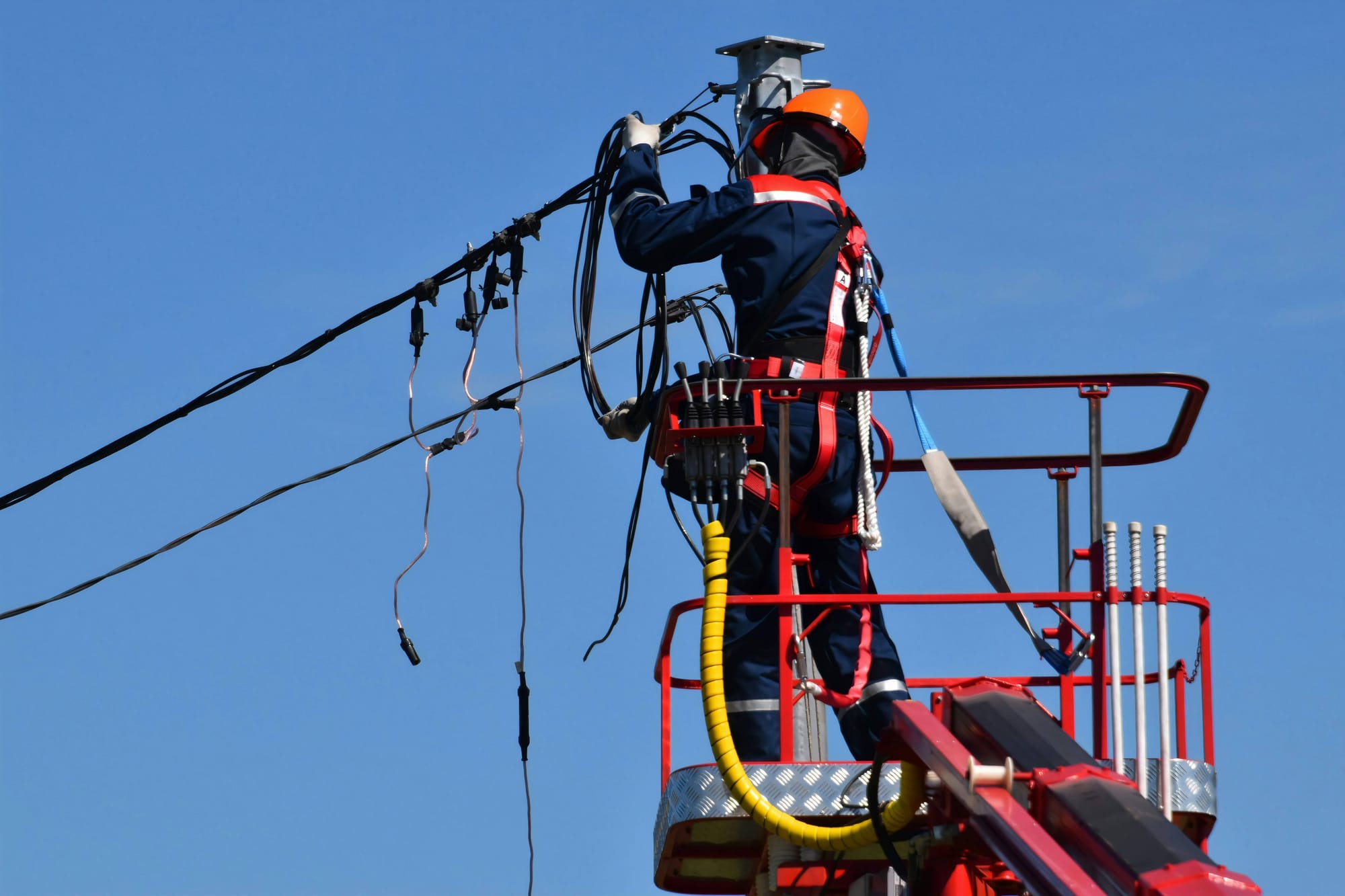 A technician on a lift platform working on overhead power lines against a clear blue sky