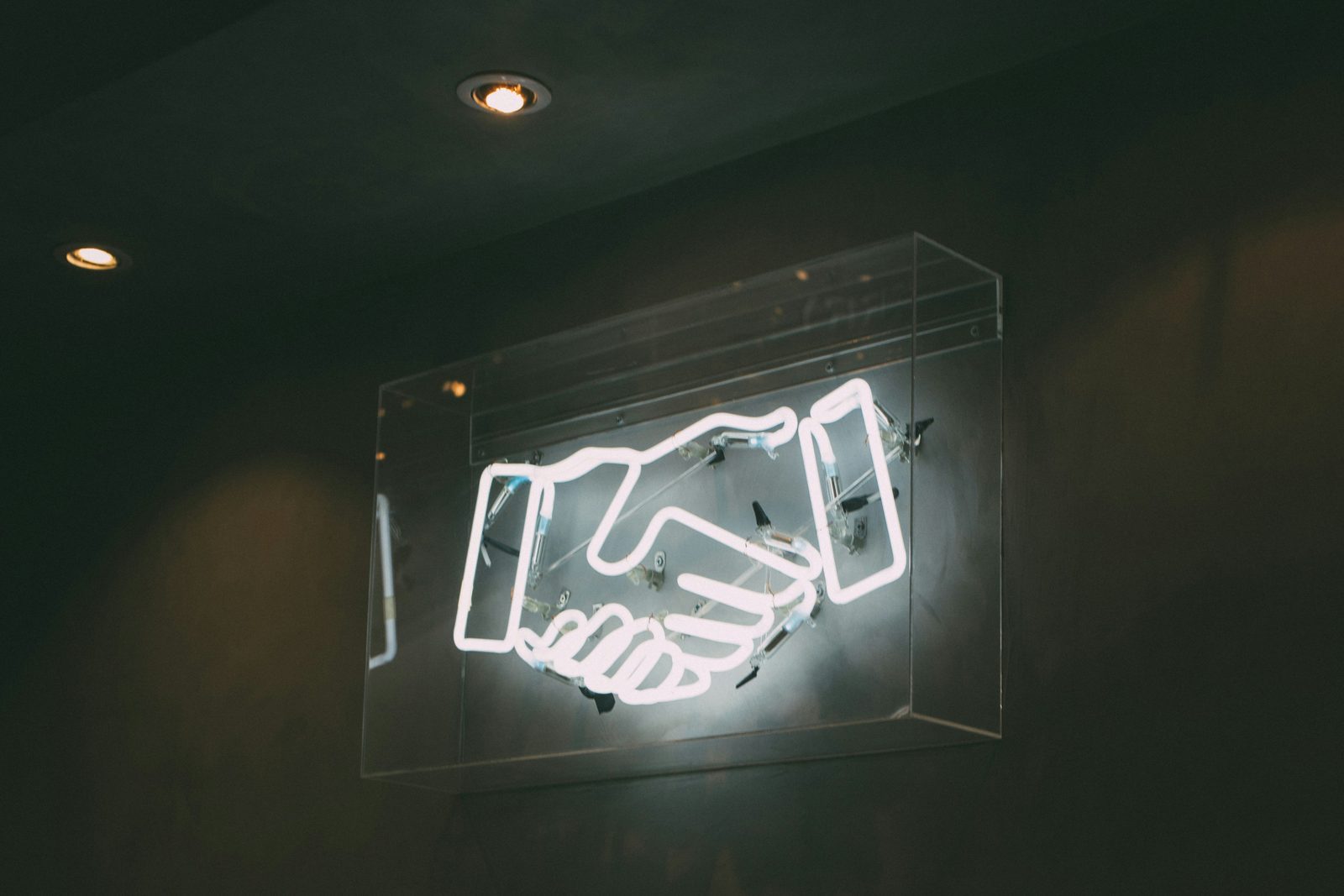 A neon sign of two hands shaking, lit inside a glass case on a dark wall