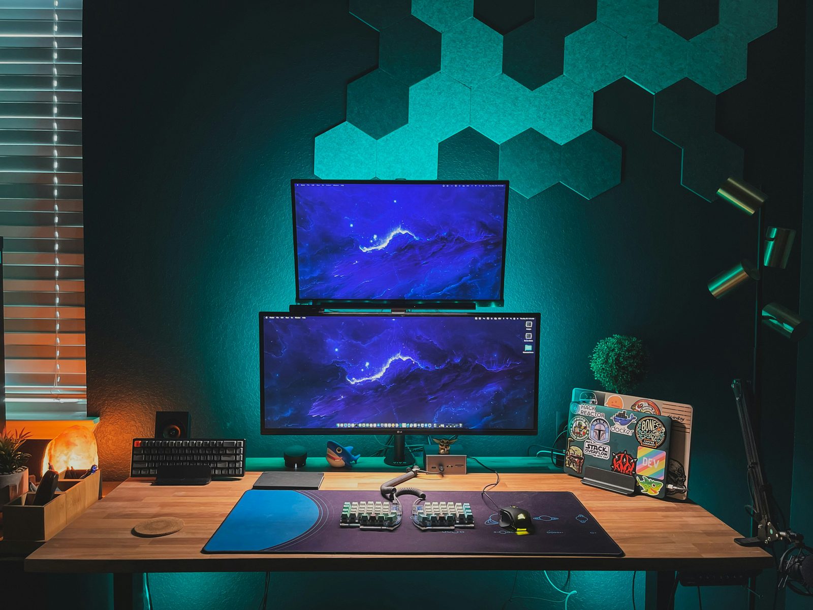 A teal-lit desk with two monitors below a hexagon acoustic panel wall