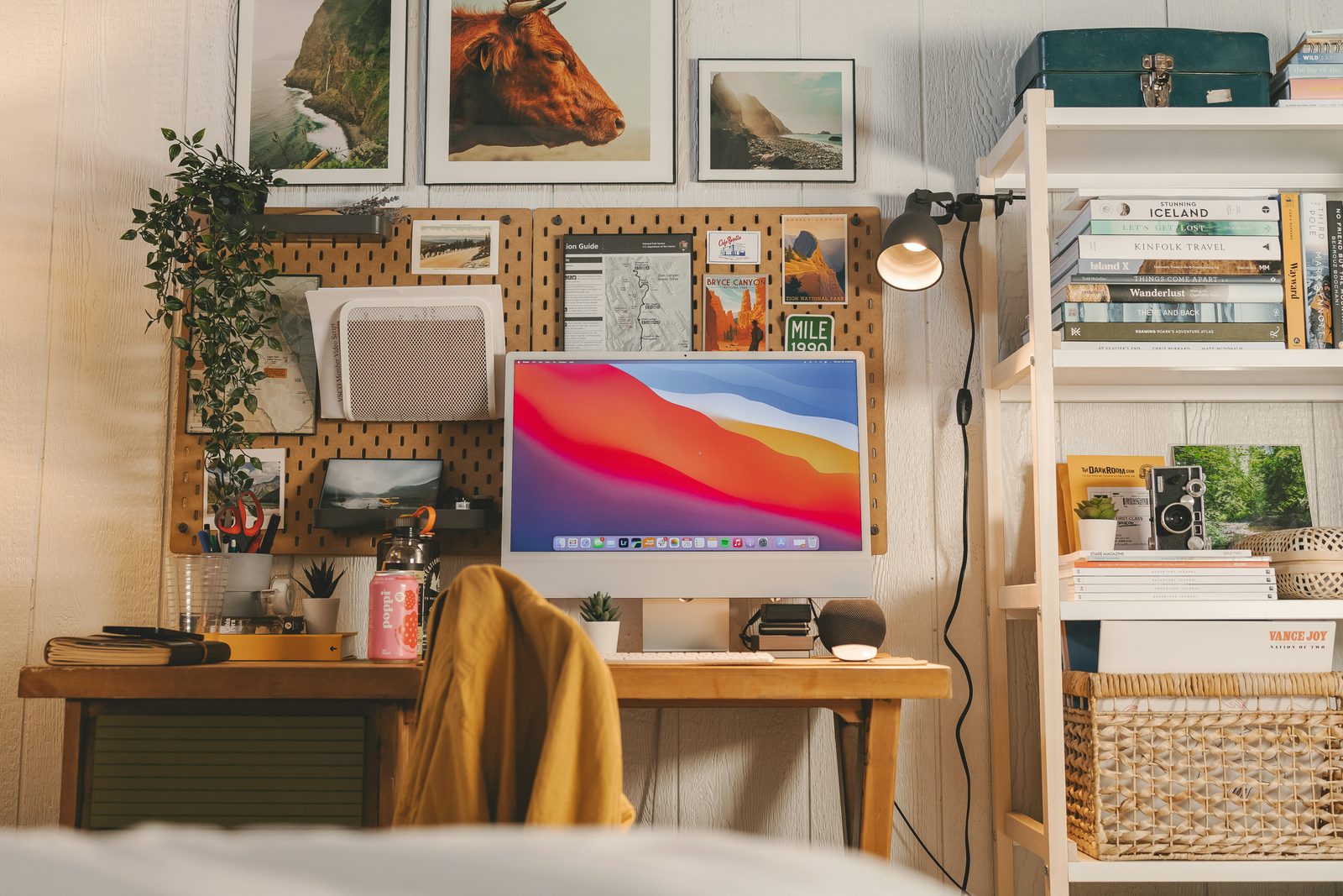 A cozy desk against a wall covered in travel photos and a pegboard, with an iMac and a bookshelf beside it