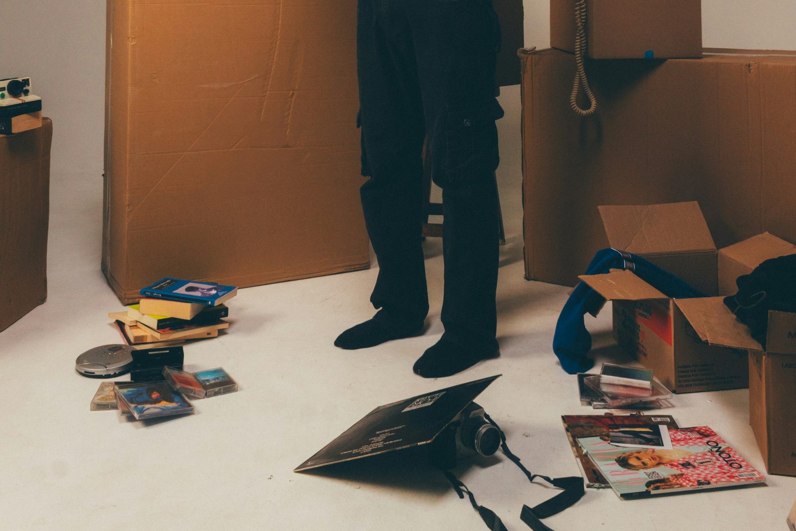 Somebody standing among moving boxes with books, cassettes and a camera spread across the floor