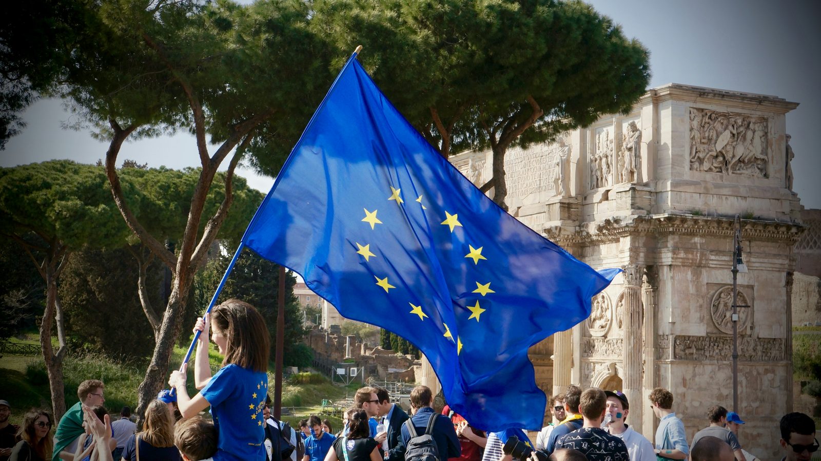 Somebody waving a large EU flag in a crowd in front of a Roman arch