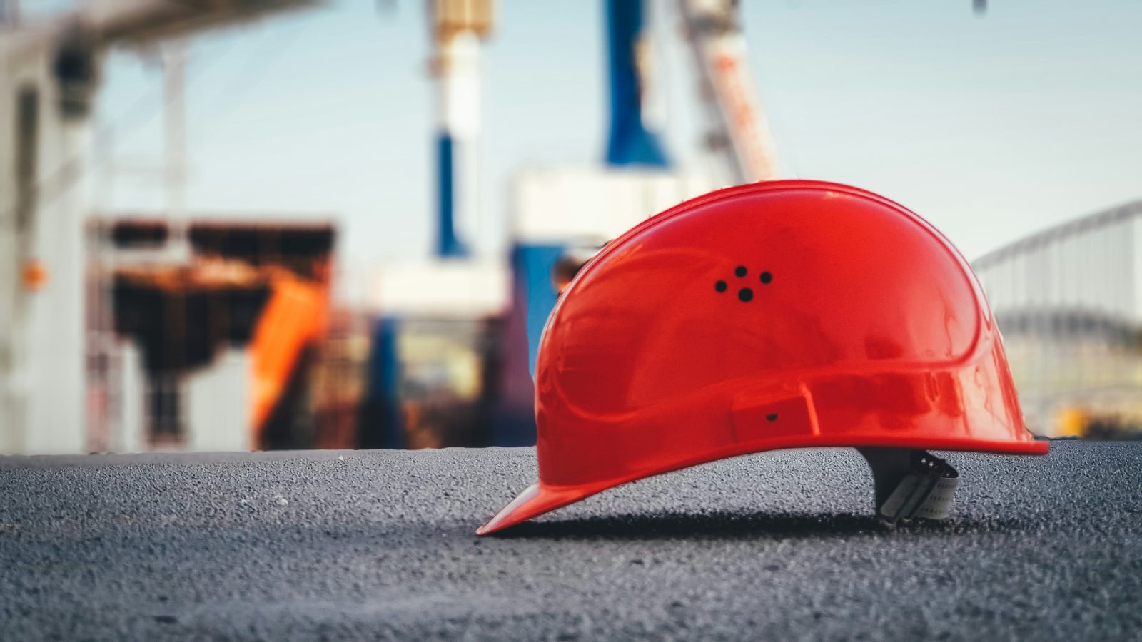 A red hard hat on the ground at a building site, cranes out of focus behind it