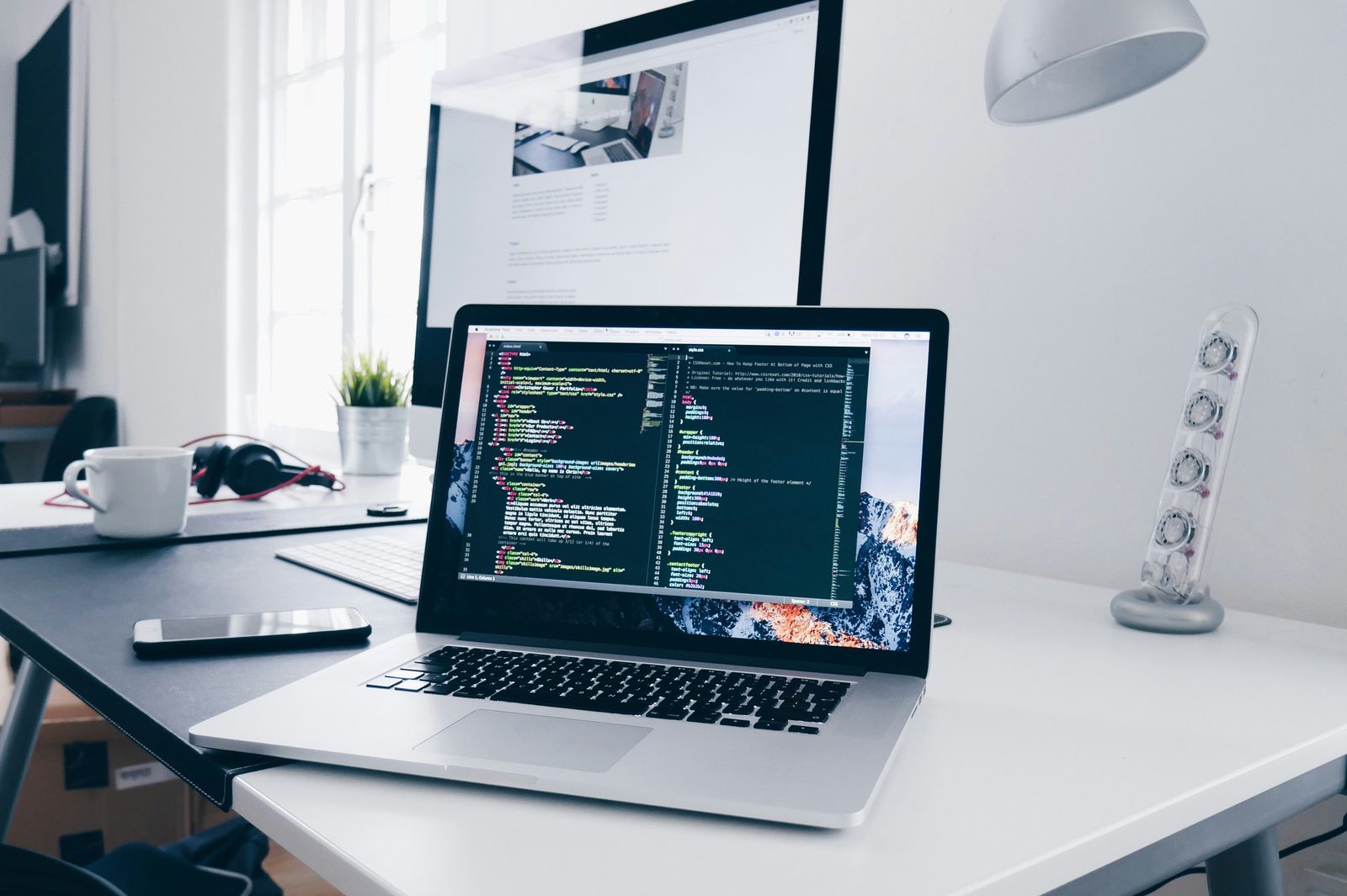 A laptop on a bright desk with a code editor open, a second screen behind it
