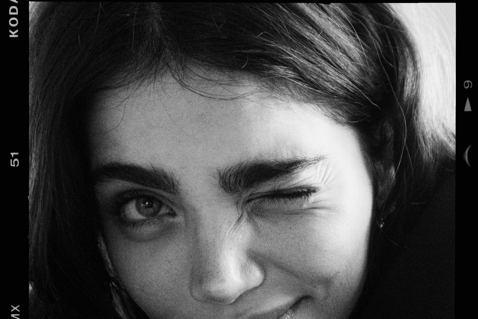 A black and white film frame of a young woman winking at the camera, chin resting on her arm