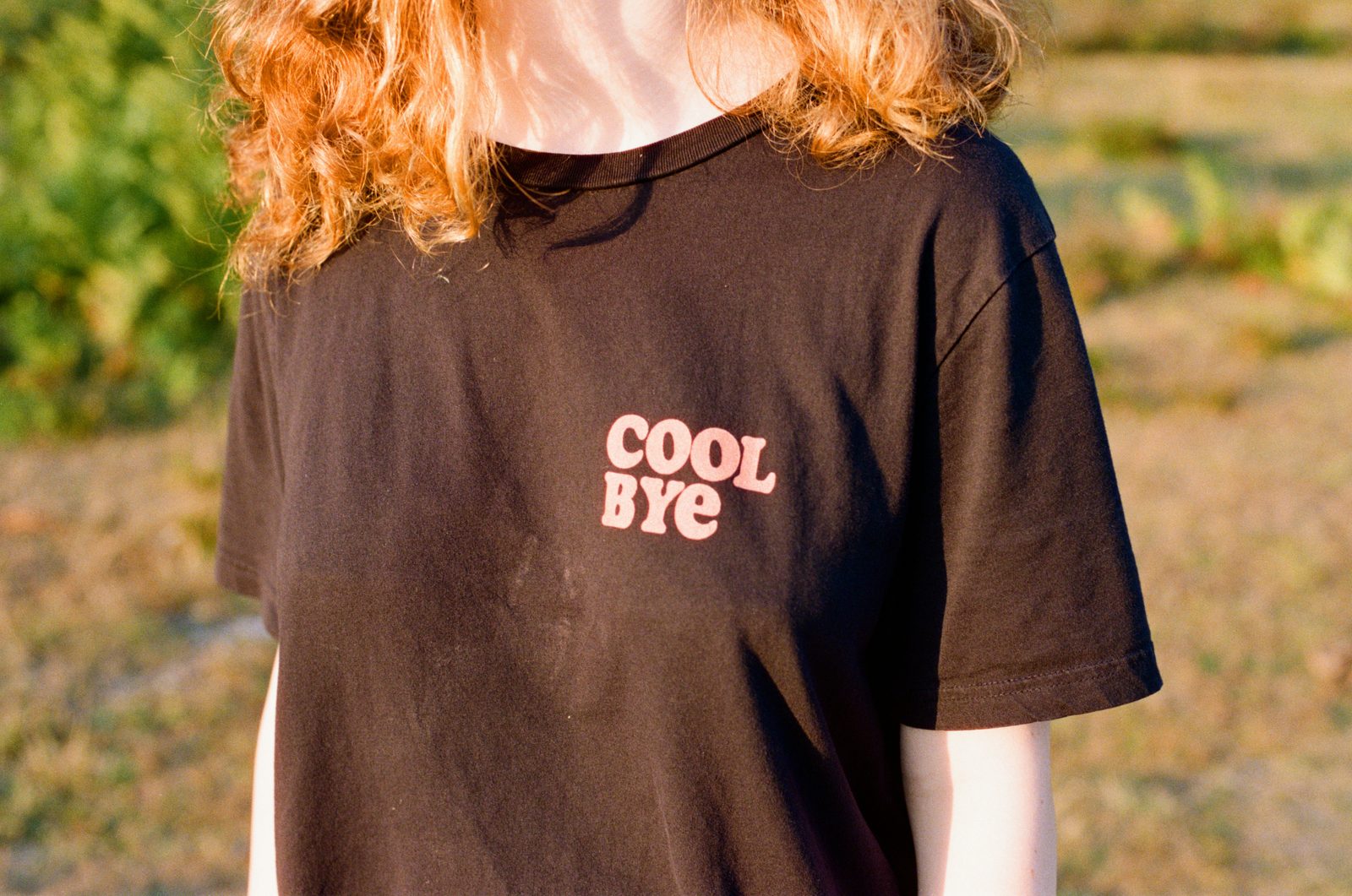 A brown t-shirt printed with the words cool bye, worn outdoors in low evening sun