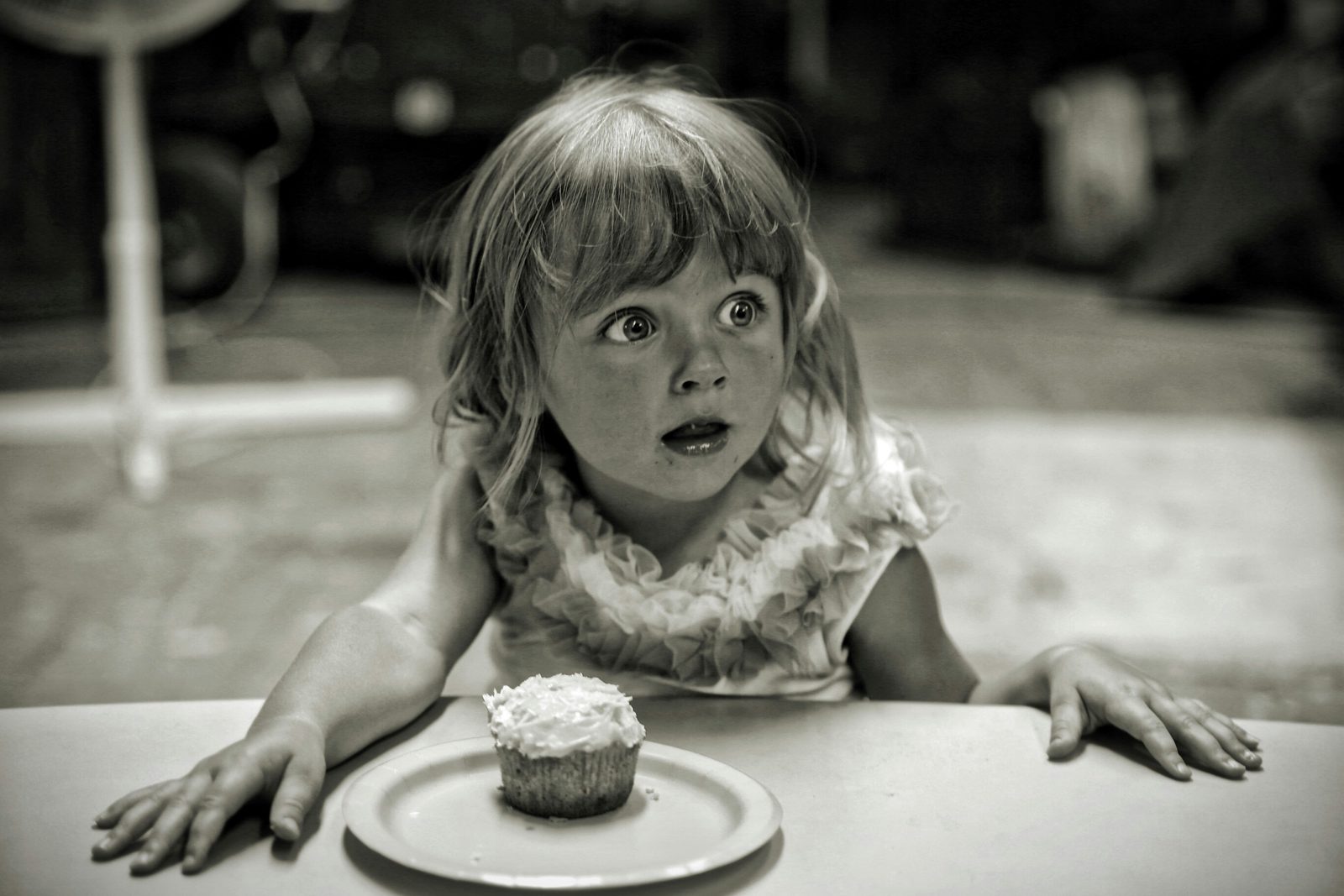 A wide-eyed girl at a table, staring past a cupcake on a paper plate