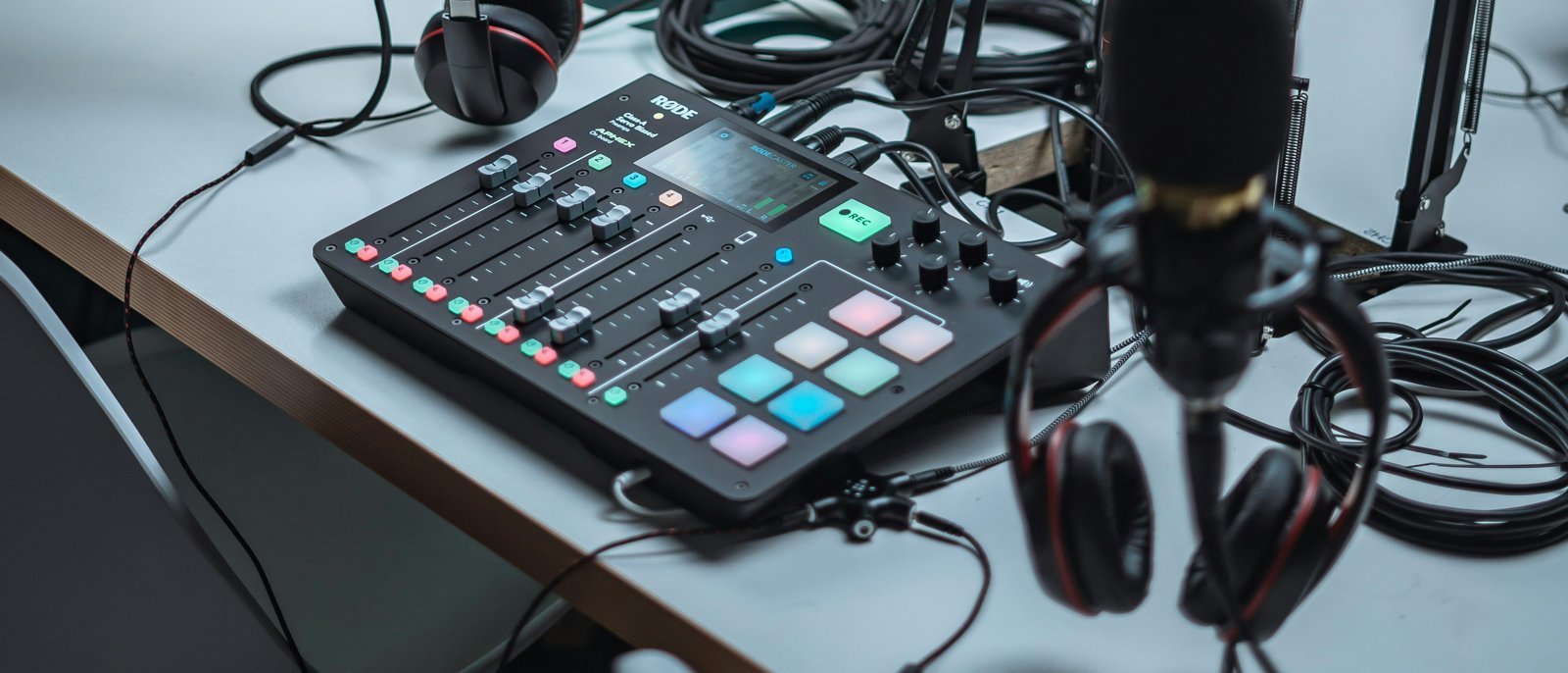 A RØDECaster podcast mixer with its pads lit up, a microphone and headphones on a studio desk