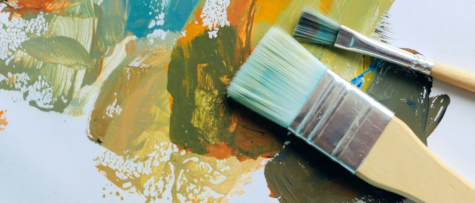 Two paintbrushes mixing blue, orange, and olive paint on a palette