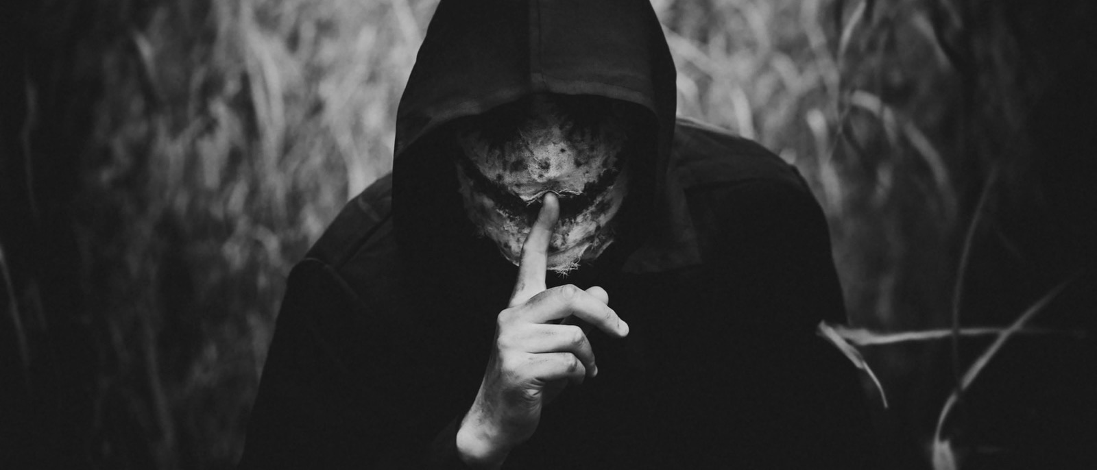 A hooded figure in a field, face obscured, holding one finger to their lips in a shushing gesture, in black and white