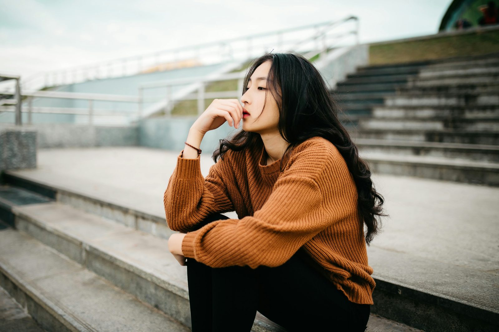 A young woman in a rust-coloured knit sweater sitting alone on wide concrete steps, chin on her hand, looking away