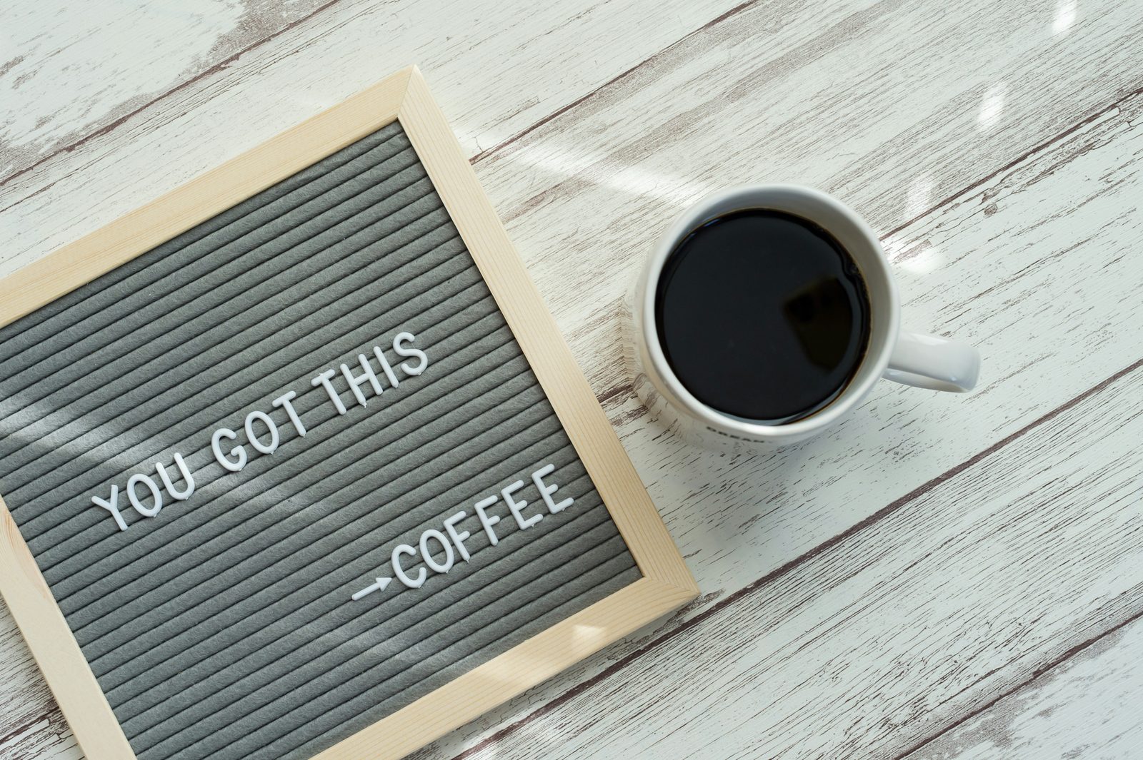 A grey letter board reading You got this, with an arrow pointing at the word coffee, next to a mug of black coffee on a whitewashed table