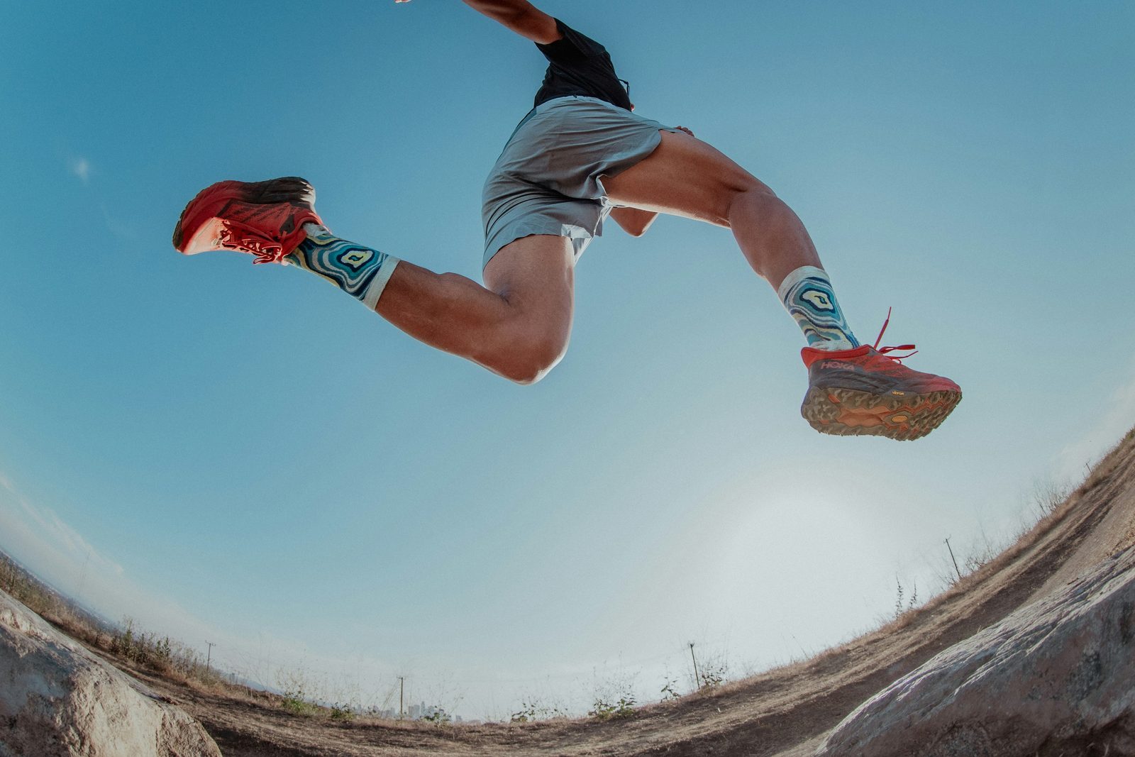 A runner in red shoes mid-jump against a blue sky, photographed from below with a fisheye lens so the ground curves beneath them
