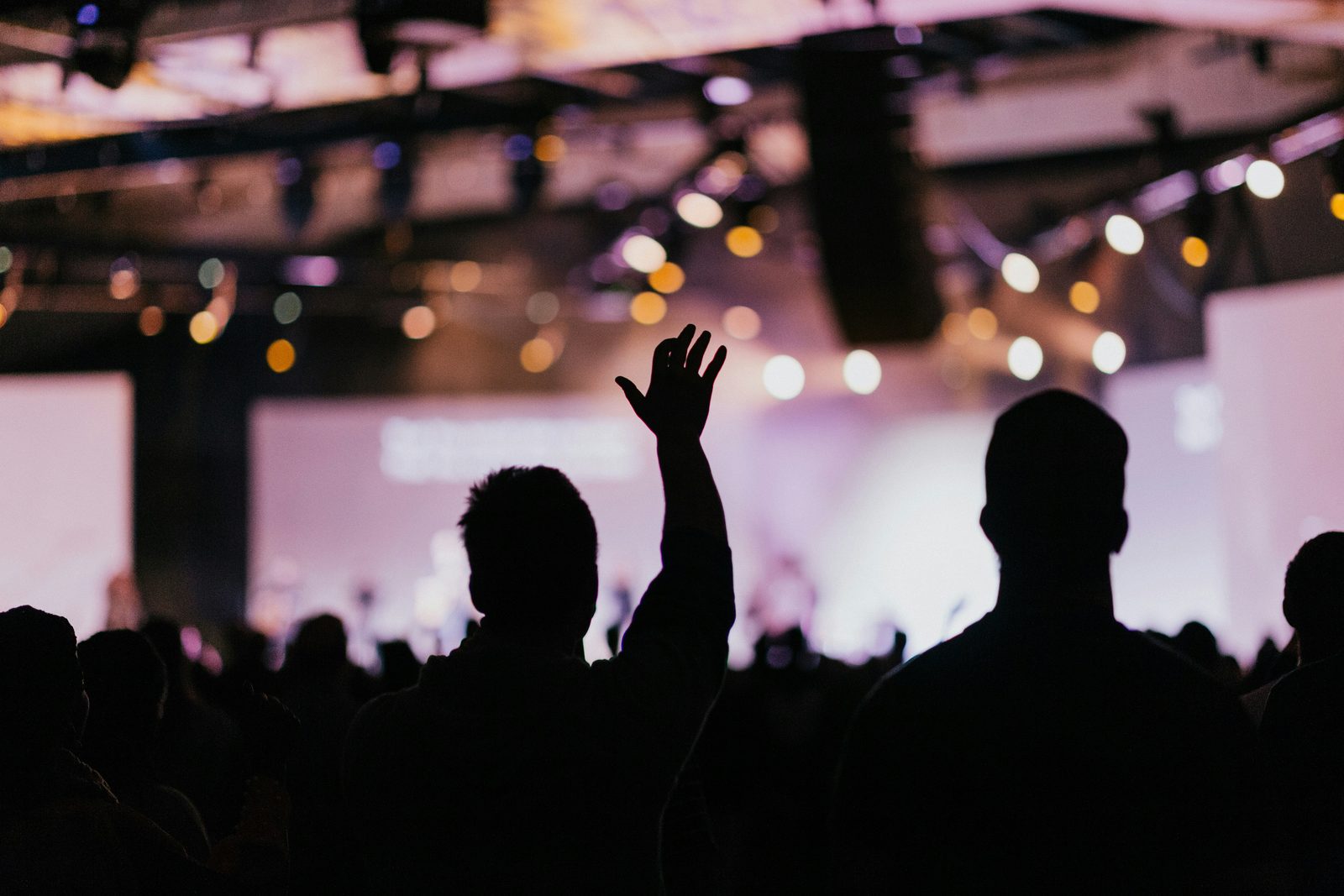 Silhouettes of an audience at a conference, one person in the middle raising a hand towards a lit stage