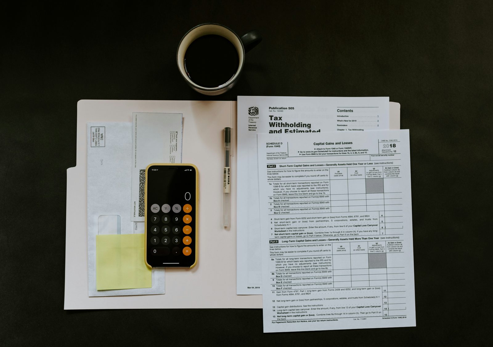 Tax forms, a calculator app on a phone, a pen and a cup of coffee laid out on a dark desk