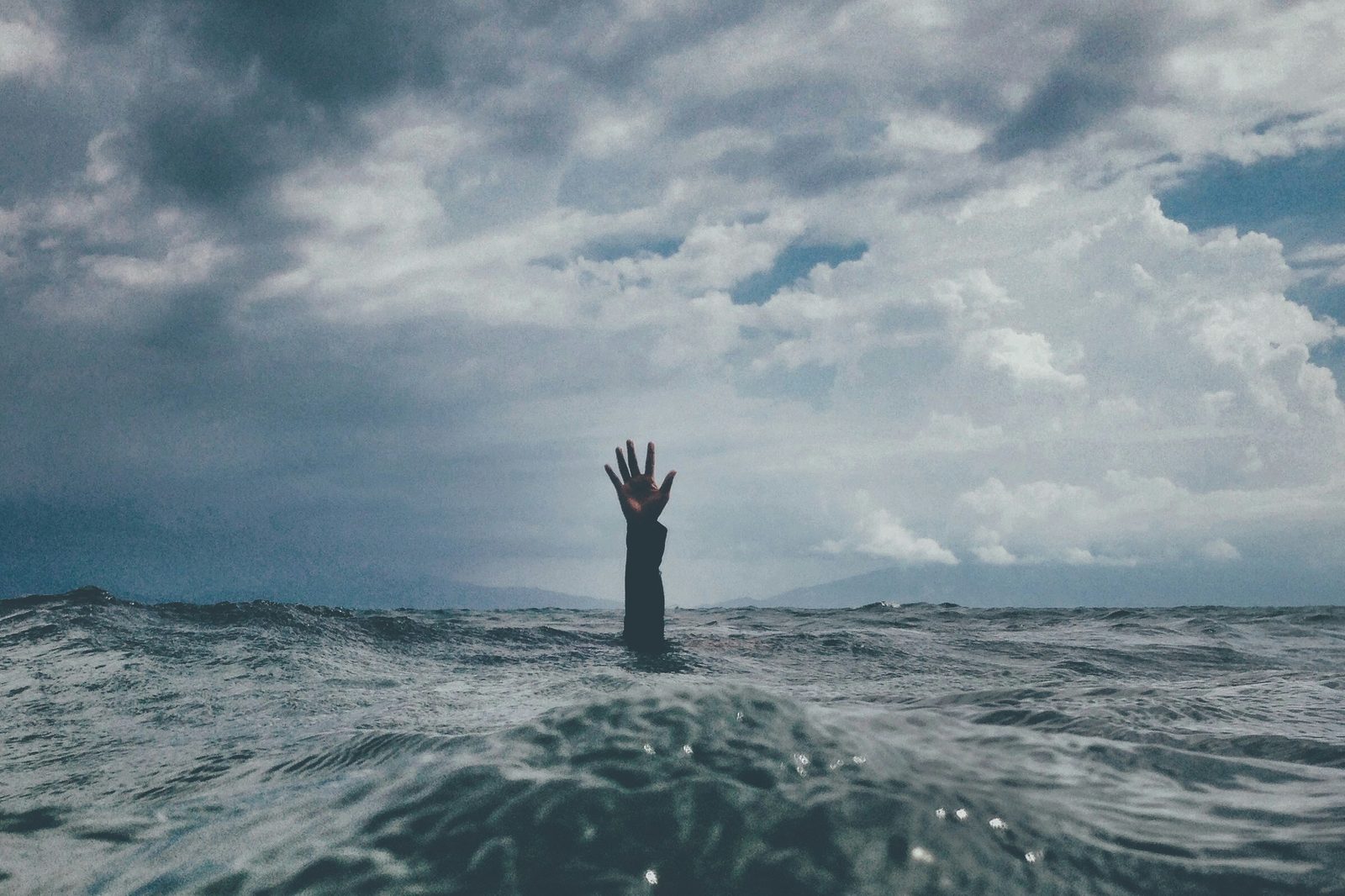 A single hand reaching up out of a rough grey sea under a cloudy sky
