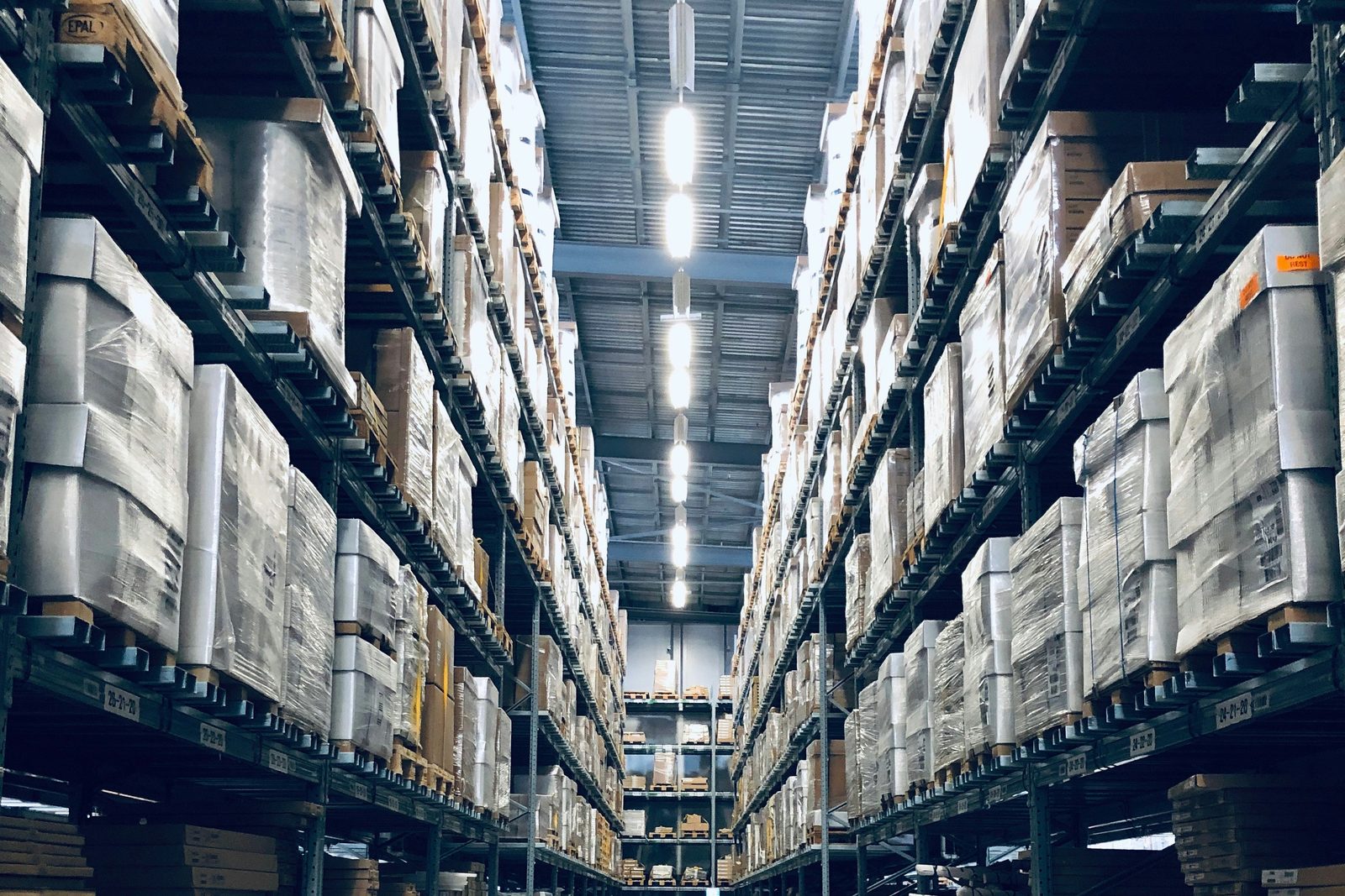 A long aisle in a warehouse, pallets shrink-wrapped and stacked high on both sides under a row of lights