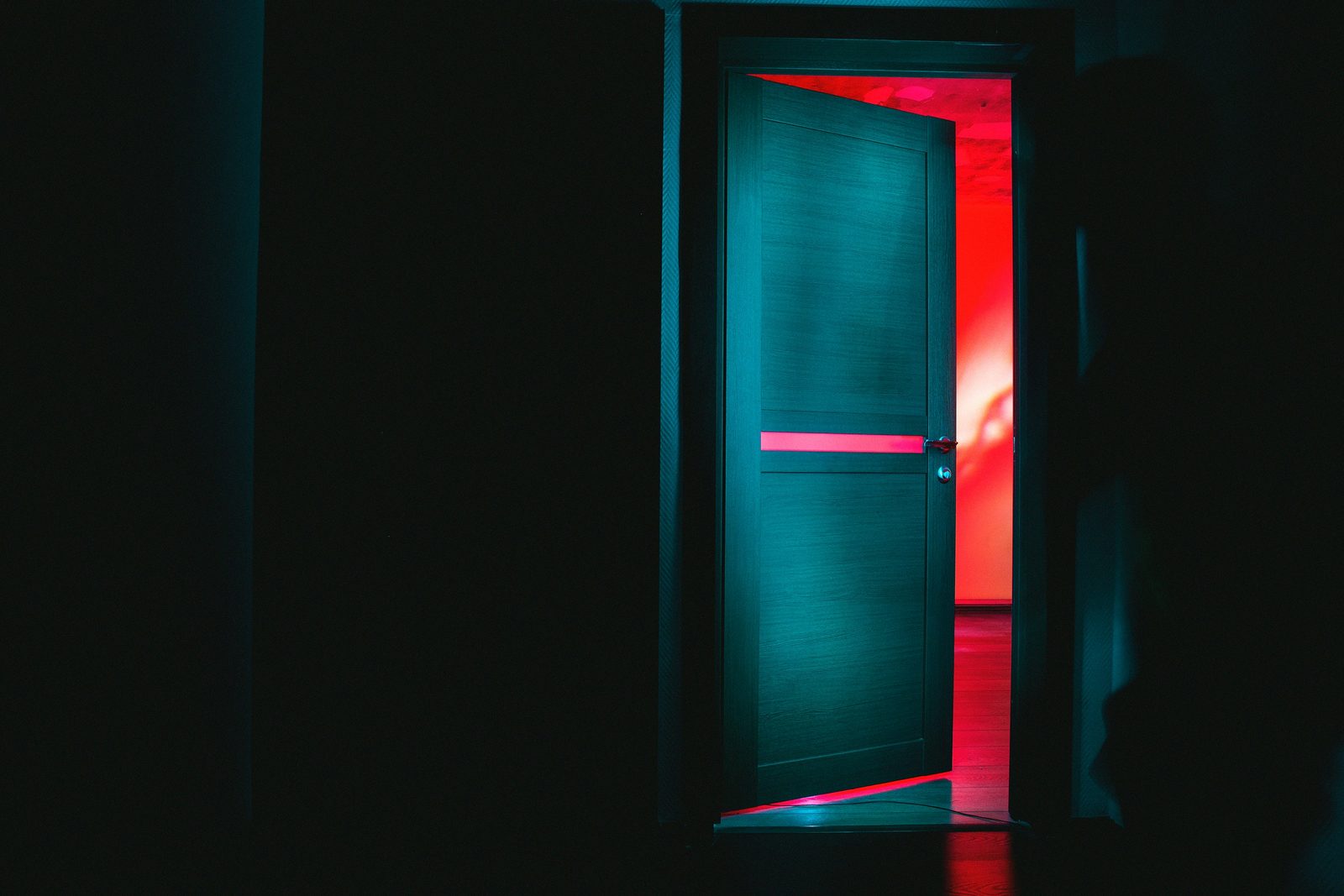 A dark hallway with a door standing ajar, red light spilling out of the room behind it