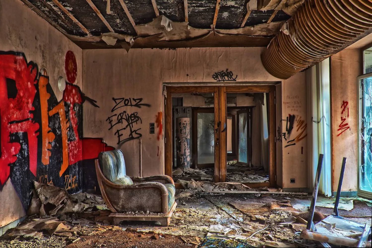 An abandoned room with a collapsed ceiling and graffiti on the walls. In the middle of the rubble a single armchair still stands upright, undamaged.