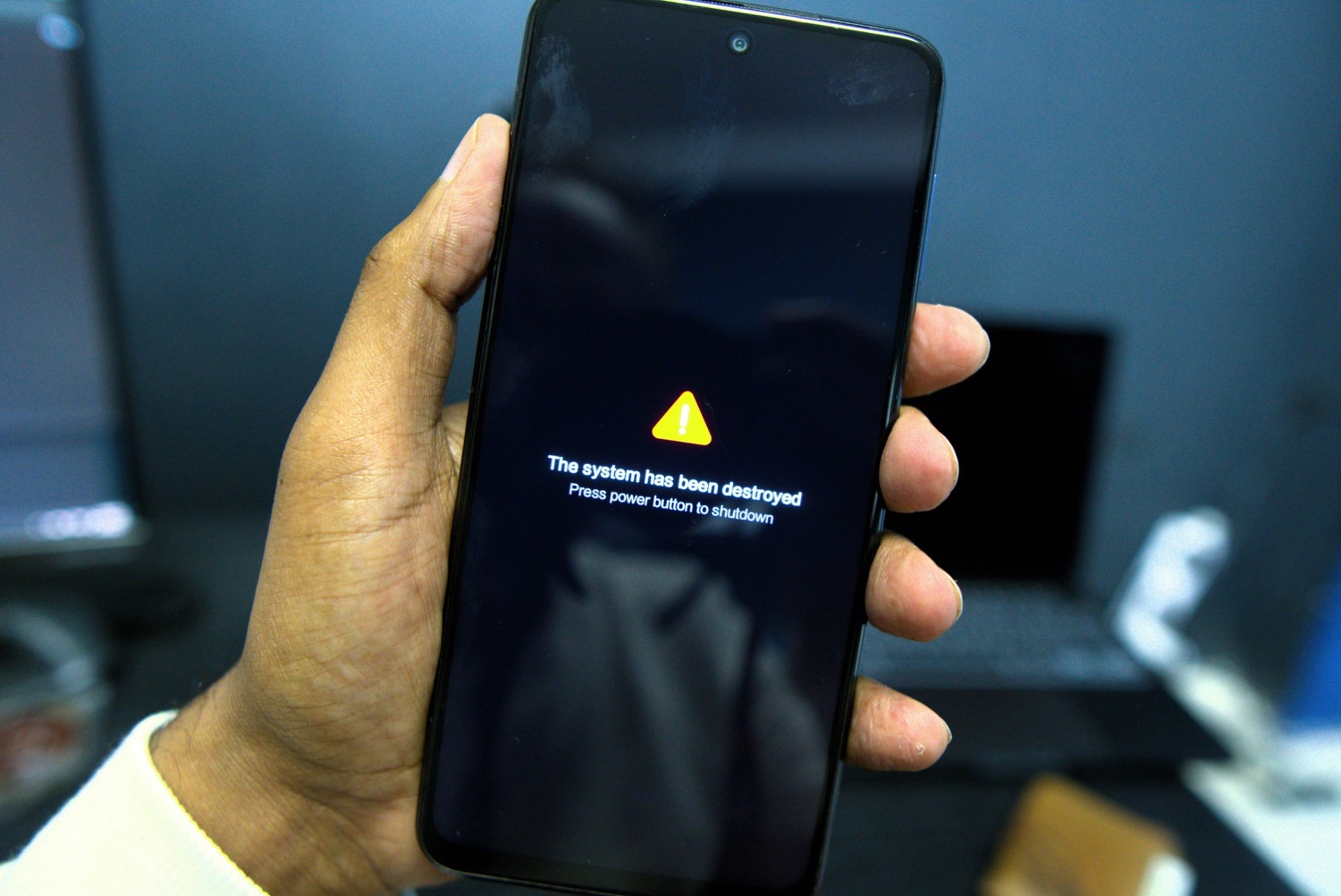 A hand holding a phone whose screen shows a yellow warning triangle above the words: the system has been destroyed, press power button to shutdown